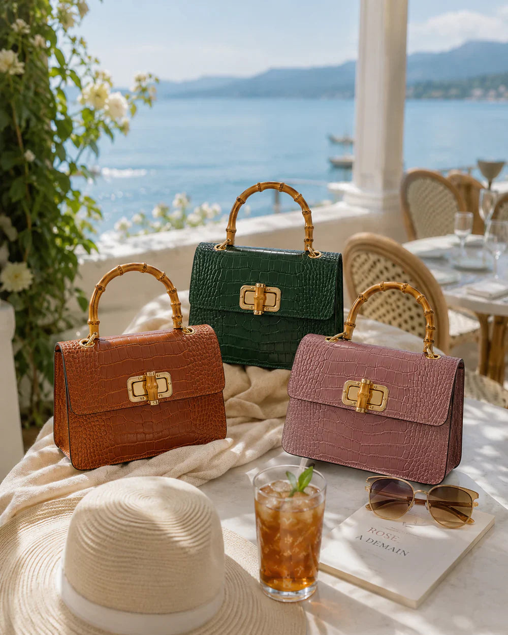 Italian croc-embossed leather bags with curved bamboo handle in dark tan, green and smoke rose, styled on a coastal terrace table